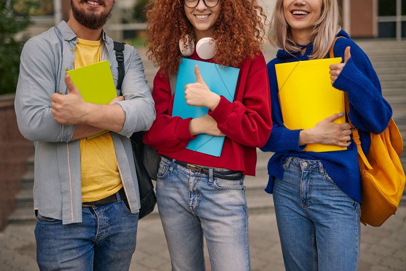 Anonymous man and women in casual clothes with colorful notepad and folders, smiling and gesturing thumbs up while standing outside university building and approving education