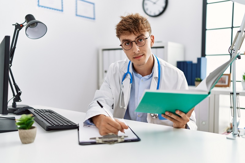 Young caucasian man wearing doctor uniform reading book at clinic